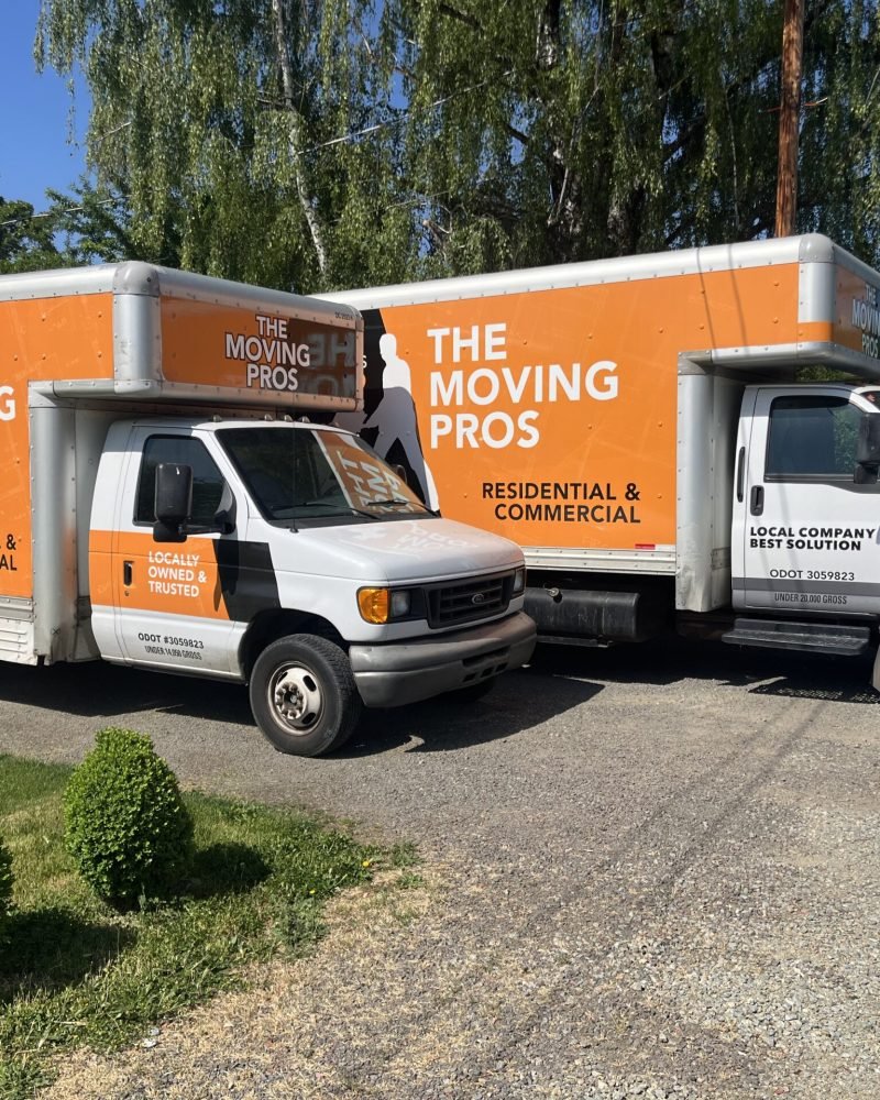 Two orange and white The Moving Pros box trucks parked side by side, branded for residential and commercial moving services in Medford, Oregon.